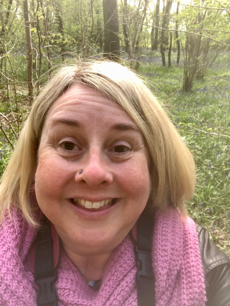 My smiling face amidst trees, I have blond bobbed hair and brown eyes, I have a purple nose stud on the left of my nose I have a pink scarf around my neck and a camera strap. Behind me the sun gives everything a pale glow there are small low trees and thicker trunks and on the ground amongst the green are swathes of blue bells