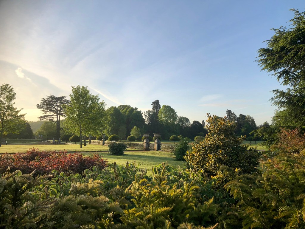 A landscape image with low shrubs and bushes in the foreground in greens, yellows and coppery purple. Beyond this is a lawn with two large urns forming a gateway, In the distance are tall trees leading to woodland. The sky is blue with fine high white clouds.  The light is misty as it is not long after dawn.