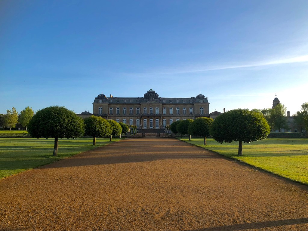 A large symmetrical stately home with steps at the front leading up to French windows. A gravel path leads up to the house flanked by small ornamental trees trimmed into a mushroom shape. The key is blue the light is hazy.  