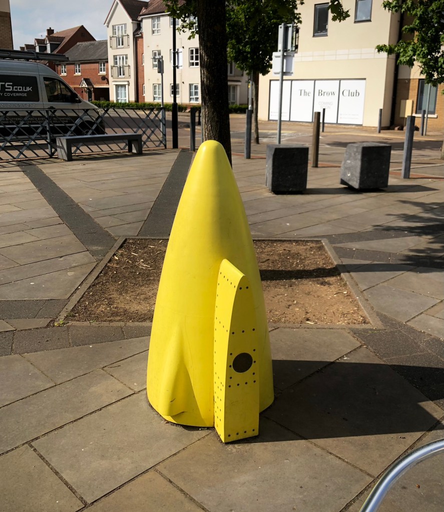 In front of a small tree is a bright yellow cone shaped monument with a wedge sticking out of it. The wedge has a black circle in the centre and black screws or rivets in it. The cone is standing in a square with benches and grey square rocks behind it. The square is surrounded by road and houses.