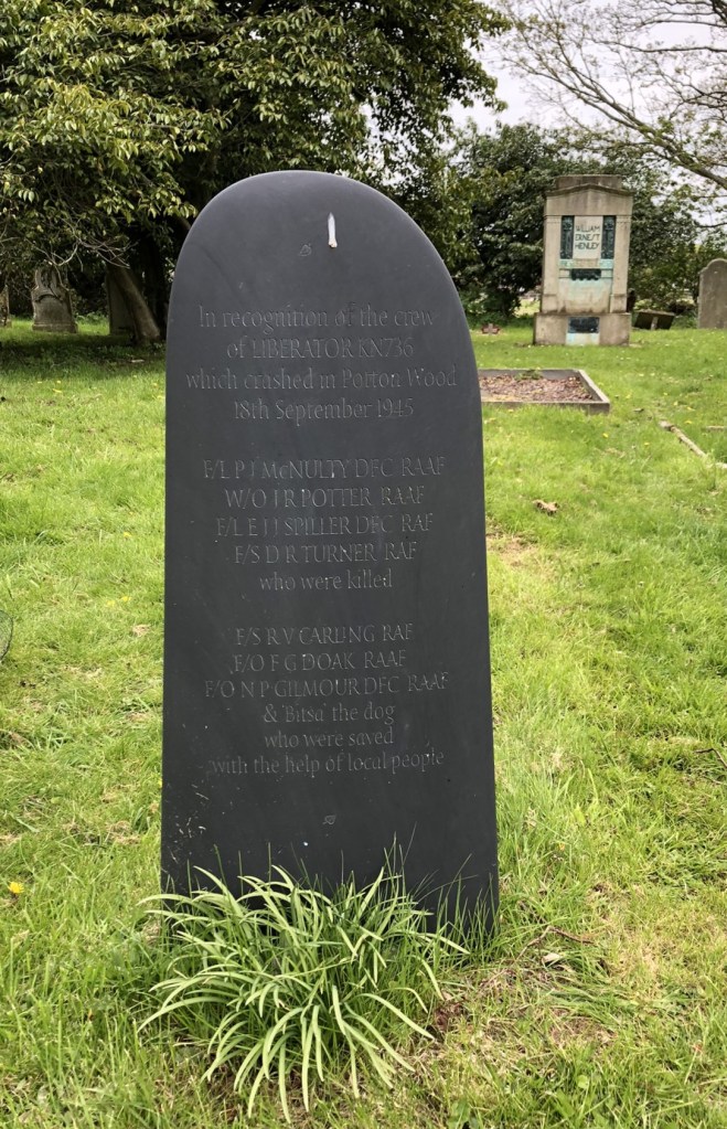 A dark grey slate wing shaped memorial. It stands in the grass of the church yards, graves are in the background along with trees.