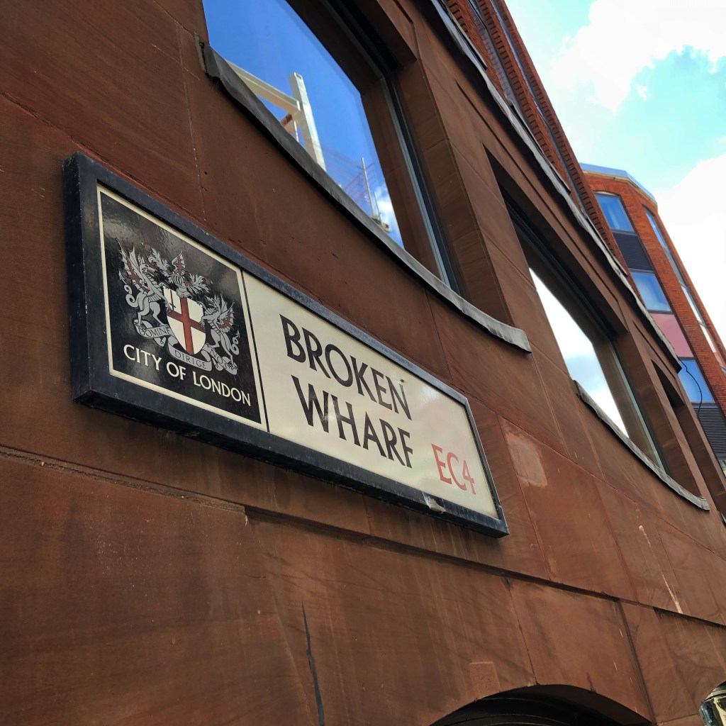 A city of London street sign with the coat of arms for the city and then Broken Wharf EC4 written on it. The sign is on a red brick building with large curved windows around it. 