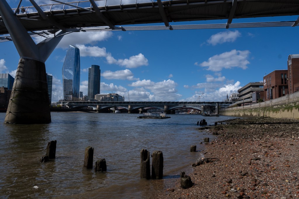 The Thames looking west from the shingle underneath the Millennium Bridge. Two sky scrapers one like mirror are to the left and Blackfriars bridge is across the river. On the right is a green weed stained wall. Old wooden posts poke out of the water at low tide like blackened teeth. 