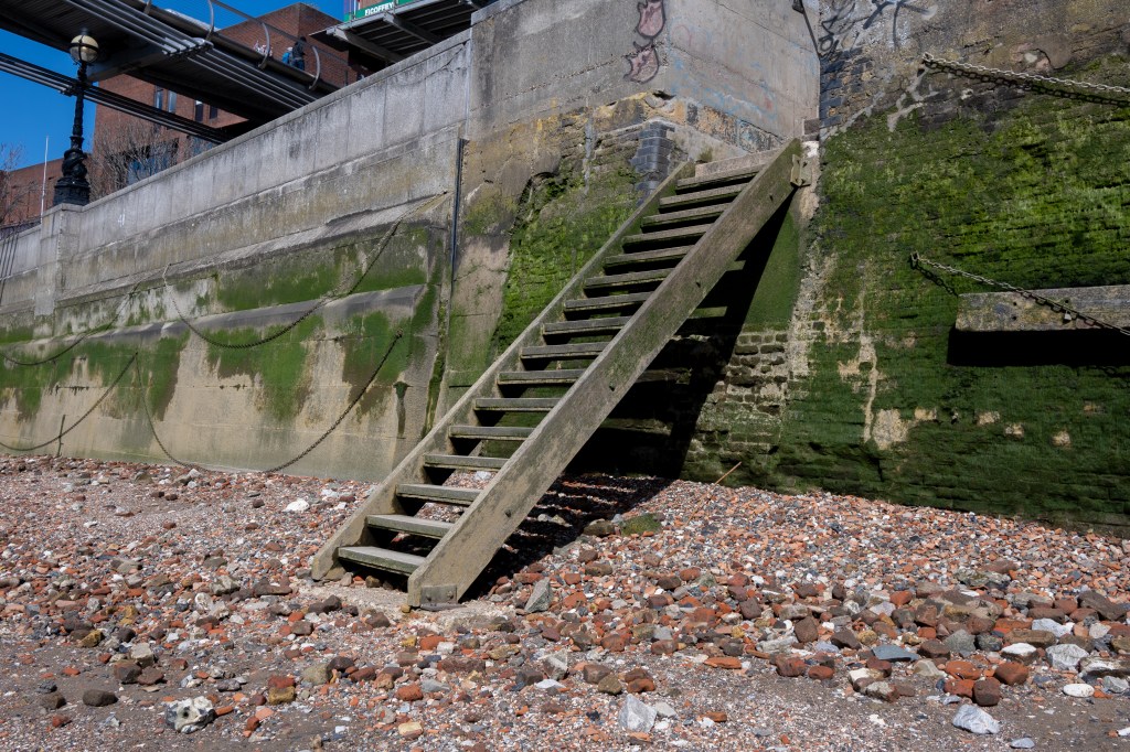 A shingle beach with a ladder like wooden stairs at a steep angle leading up onto a green weed stained wall which has chains tacked along it. The edge of the Millennium bridge is the top left hand corner. 