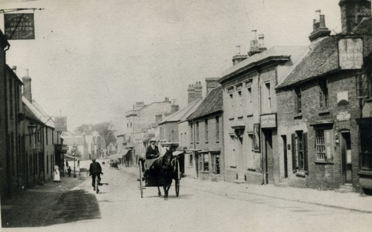 A black and white photograph of a wide street with a horse and cart in the centre coming towards us. Either side of the road are old buildings the one nearest on the right has a sign saying Golden Eagle on it. 