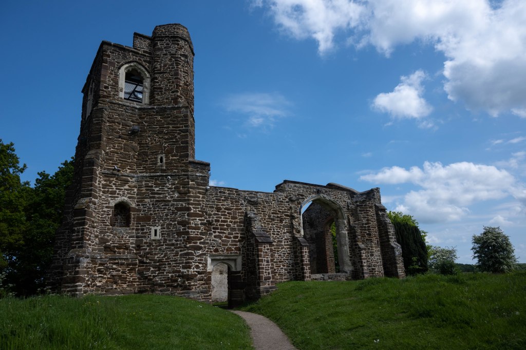 The church made of a brown and grey stone, it has no roof and the bell tower is ragged at the top a stair case can be glimpsed through the top window of the tower. There is a doorway into the church and then large arched windows at the side and end of the building. The sky is blue above with some white clouds a may blossom bush is on the far right of the picture and the grounds around the church are long grass and wild flowers. 