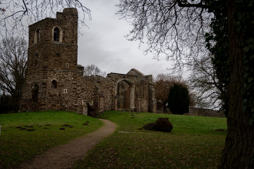 A ruined church no roof or glass in the windows, a brown and cream colour with a brown path snaking up to it's open door. There is grass around the ruin and mole hills in the foreground on the right is an ivy covered tree. 