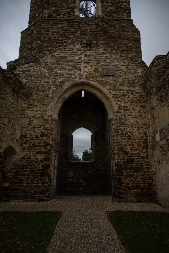 Looking into the tower from inside the church, There is a window in the tower facing on to trees and sky and a gravel path going into the tower. As the square tower rises there is another window showing the interior spiral staircase and cage at the top of the tower. The image is dark and oppressive.