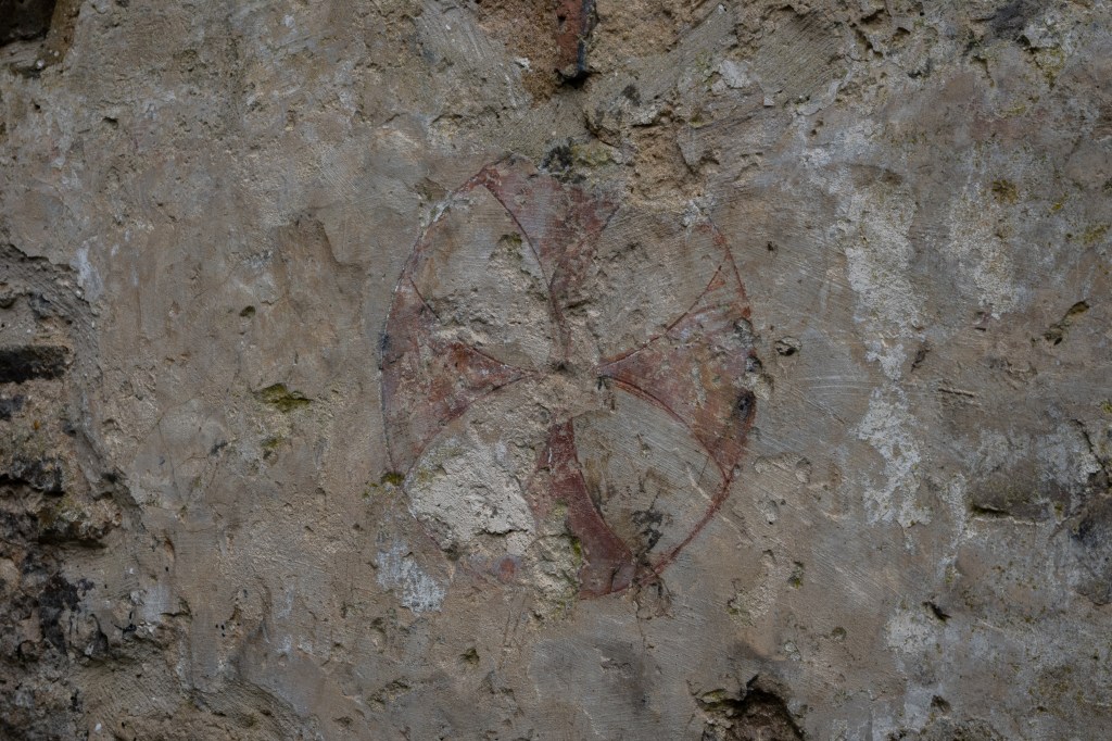 Mottled plaster with lichen and moss growing in breaks in it. In the centre a faint red Maltese cross within a red circle.