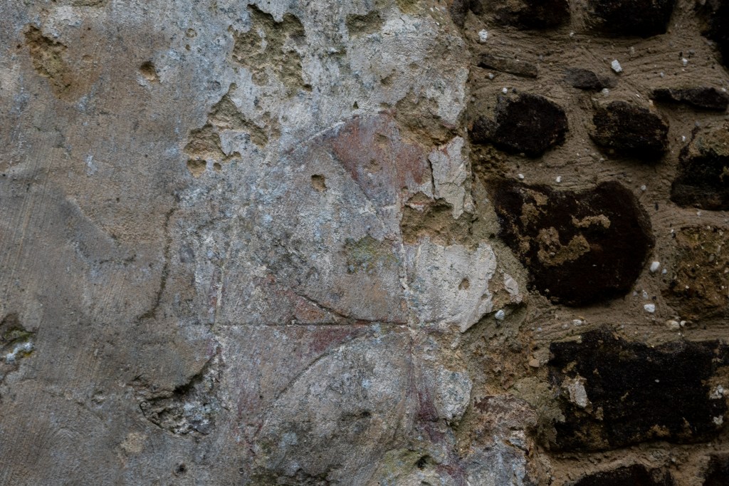 A partial Maltese Cross faintly painted on to plaster which is breaking away meaning the right hand side of the cross is missing. Next to the paster are dark stones in gravelly mortar. 