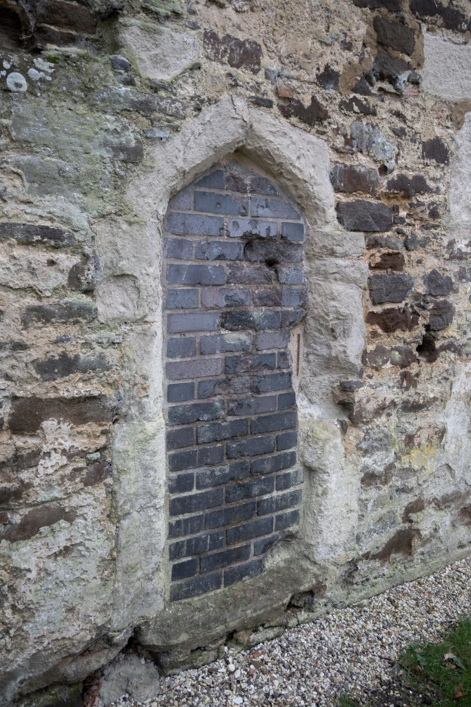 A small arch way with a step which is bricked up in more modern grey brick inside the church. below it is gravel and grass. 