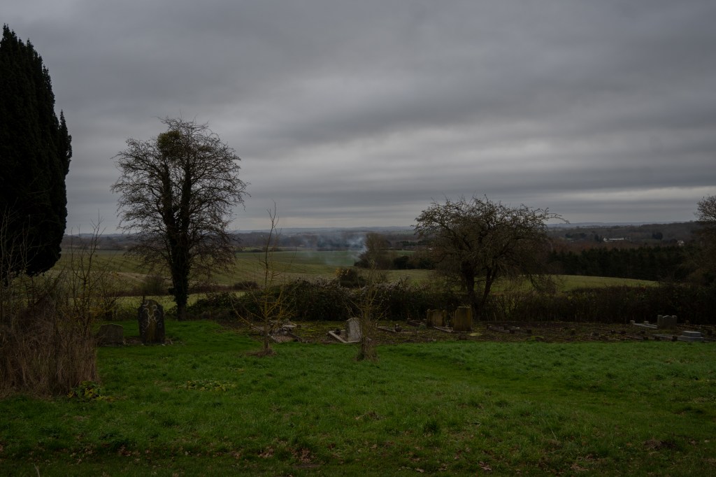 Grass and grave stones against a low hedge then fields sweeping into the distance. Smoke is rising from below and the sky is grey streaked with white. There are winter trees in the middle distance. 