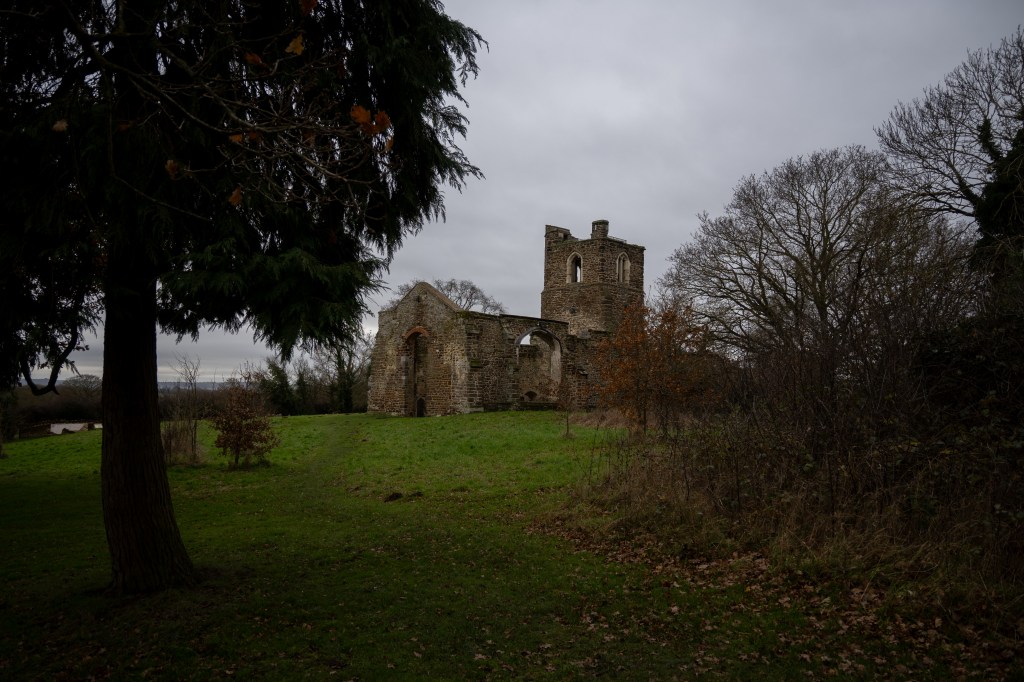 The church glimpsed between a large ever green tree on the left and low skeletal winter buses on the right. The door way to the church is open and a dark interior doorway and then mottled paler markings above it can give the impression of a figure in the door way. You may also see other figures on the outside of the door in paler stones. 