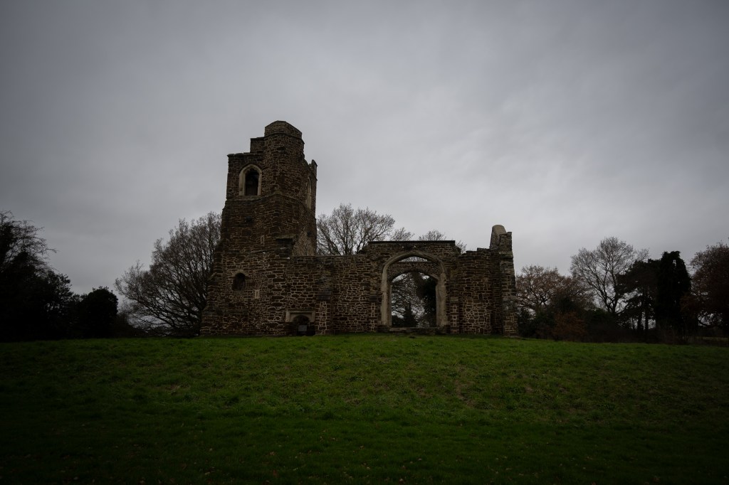 The ruined church taken from slightly below it with the grass mound it sits on rising. The empty windows line up so that you can see straight through both of them to the winter trees beyond. The square tower is jagged. It is surrounded by winter trees and the sky above is grey with white clouds daubed across it like finger prints. 