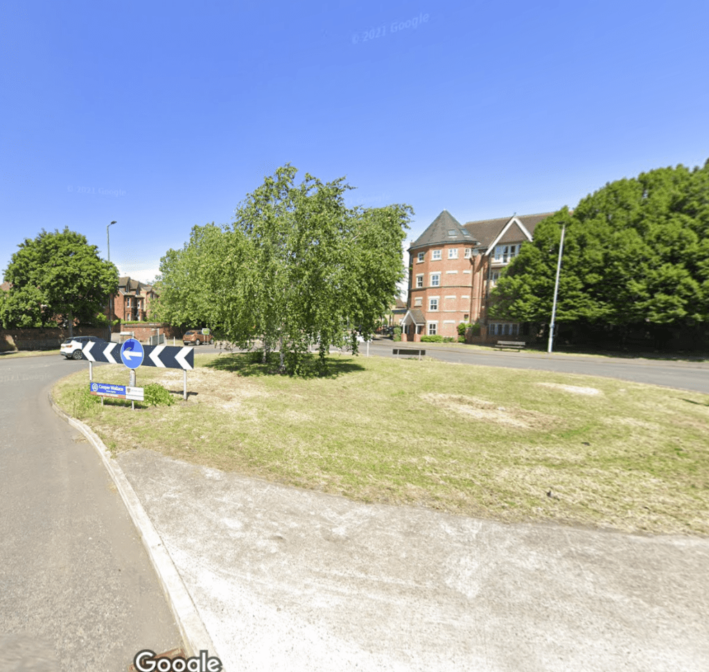 A grass covered roundabout with some small trees on it, in leaf. A tall red brick building with a tower is to the right behind the roundabout next to some bushy trees. There is a bench below the trees facing the roundabout. On the left are trees and some Victorian terraced houses. The sky is bright blue.