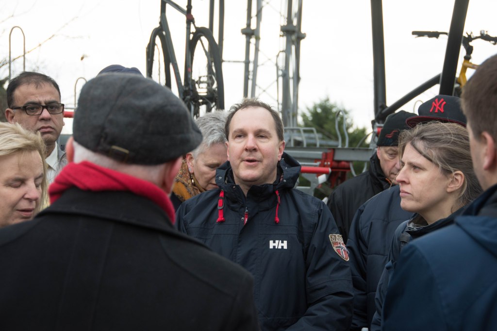 In the centre of the image a man of around 50 with short brown hair wearing a dark anorak with red toggles is speaking. Surrounding him are many people their heads and shoulders in shot. Behind them are bike racks and bikes and white winter sky.