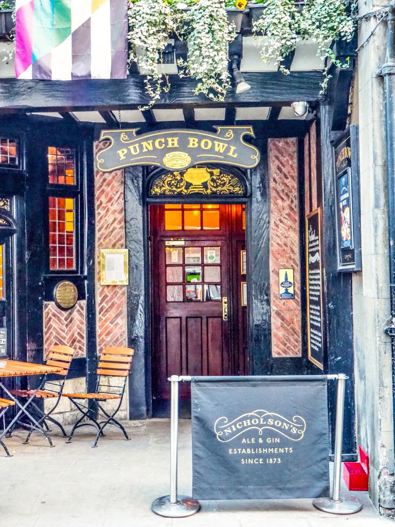 The door to an old pub. Above are wood beams against white, the door is dark brown and golden light shines through the windows. The sign above the door reads Punch Bowl. 