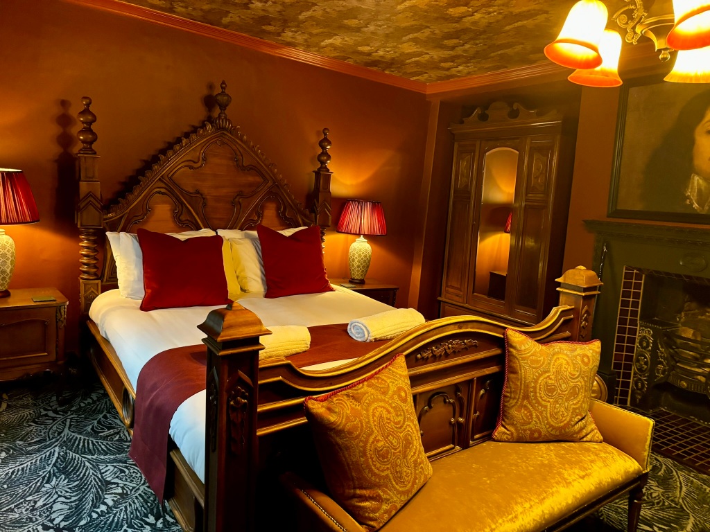 A large double bed with red cushions and the walls of the room are red as are the lamp shades. At the end of the bed there is a gold settee with cushions. in the right hand corner is an ornate wardrobe. There is a large tiled fireplace. 