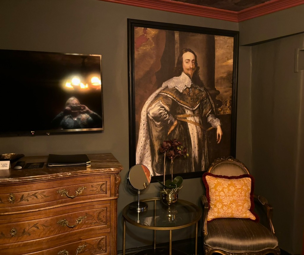 The corner of a bedroom with a fancy silk chair with gold cushion next to a round glass table. on the left there's a large chest of draws and TV above it. I am reflected in the TV taking the photograph. There is a large painting of Charles I in his silk robes. 