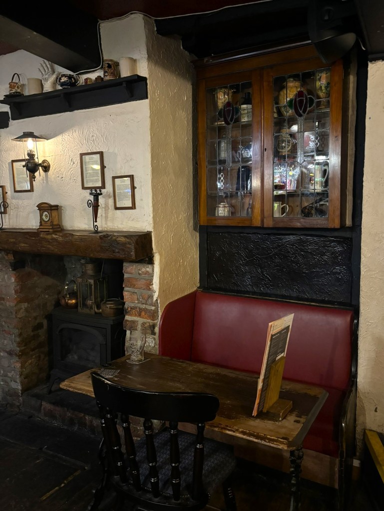 A red settle like seat at a wood table with an old glass fronted cabinet above it. On the left of the table is an old brick fireplace with rustic wooden mantle piece. There is a dark wood shelf above that with an assortment of odd ornaments.