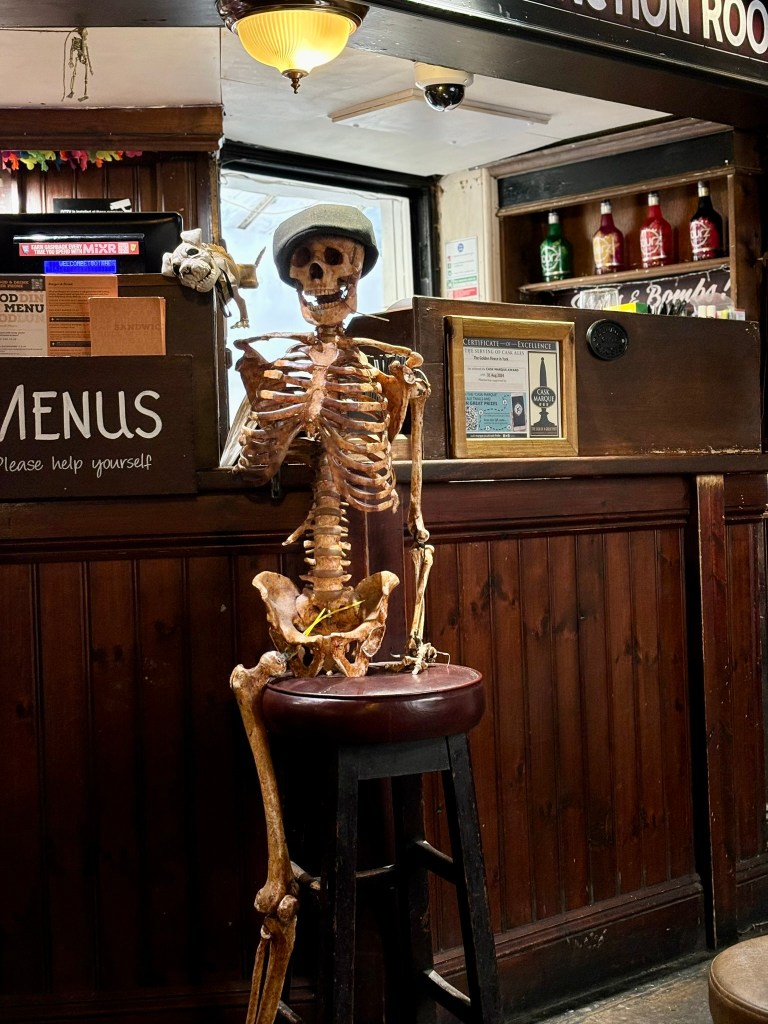 A human skeleton wearing a flat cap sitting on a bar stool at an old wodden bar.