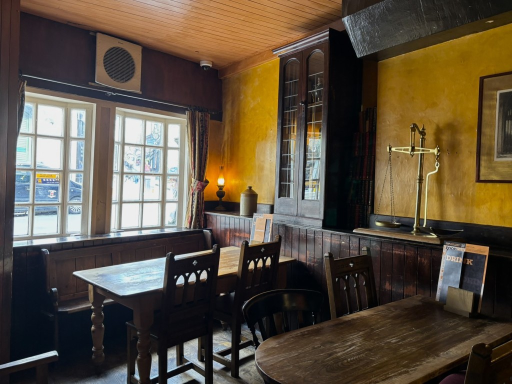 A plain bar of wooden settles and tables and chairs. The walls are a mustard yellow the ceiling has wooden boards. A large set of gold scales are on a wooden shelf next to an old wooden cabinet.  