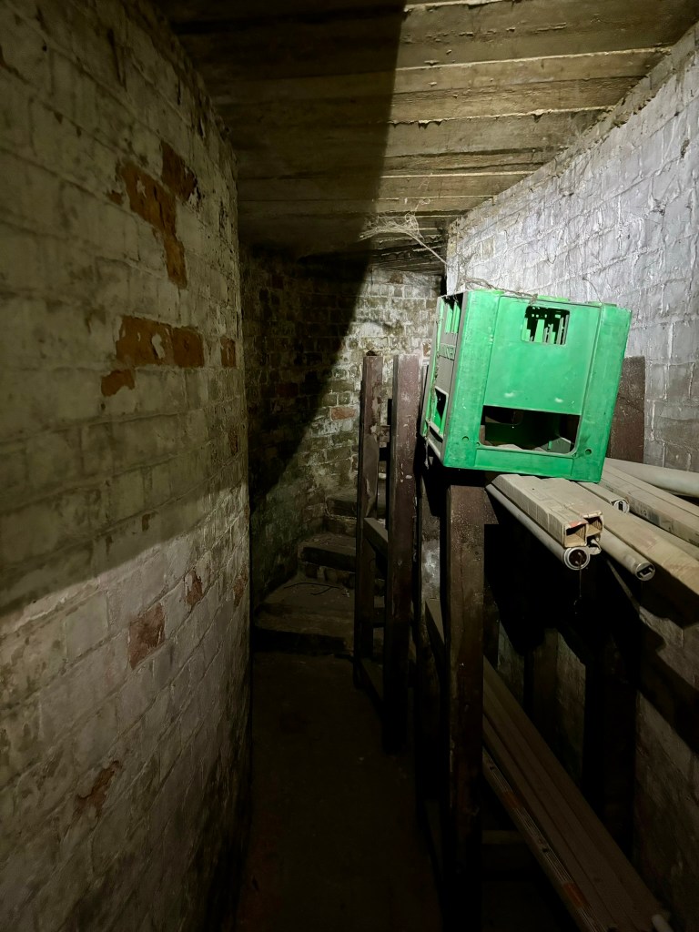 On the left a curved brick wall. Above a cobwebbed ceiling, on the right a white washed brick wall with wooden frame and beer crate stacked up. At the end of the curved passage are a few steps 