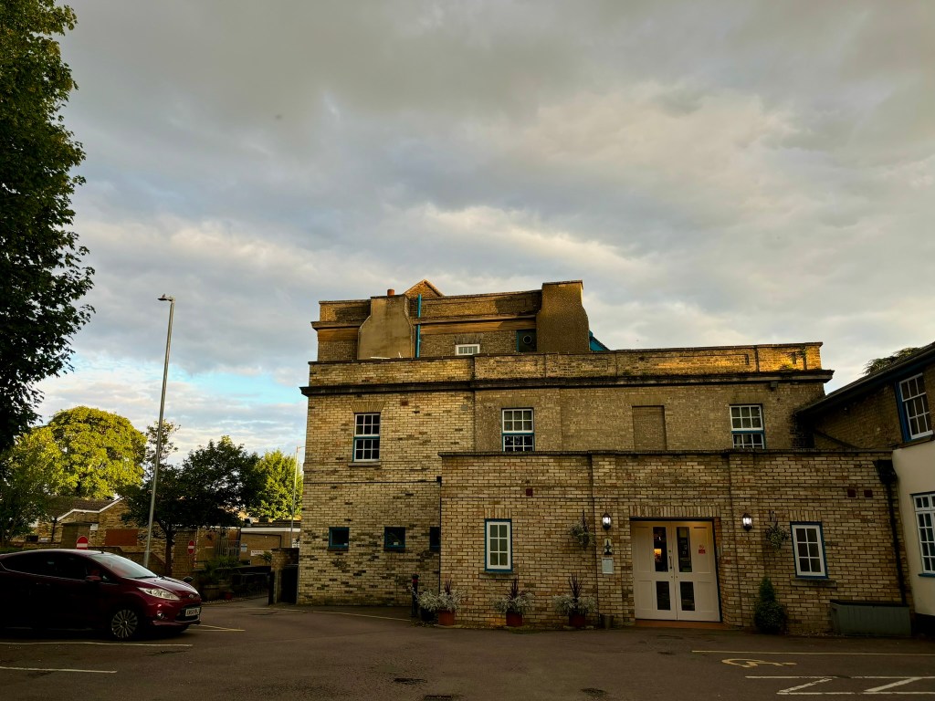 A squre pale brick building of different levels and heights. There are large chimneys and a kind of brick parapet. There are long narrow windows one of which is bricked up and wide white doors. The sky is grey with some late evening sun glinting off the building. 