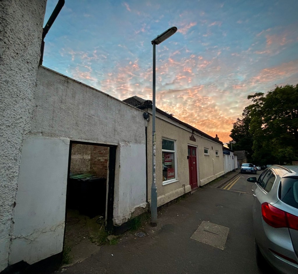The Labour Hall at sunset, through an open gateway the end of the labour hall is visible with a clear difference in the colours of the brick showing where the old back entrance to the schoolroom was accessed from Camden House. The building is long and low with two high up narrow windows. There is a more modern window next to the old red door. The building beyond it is also a low one story building of a brighter white. The sky above is dappled with orange clouds.