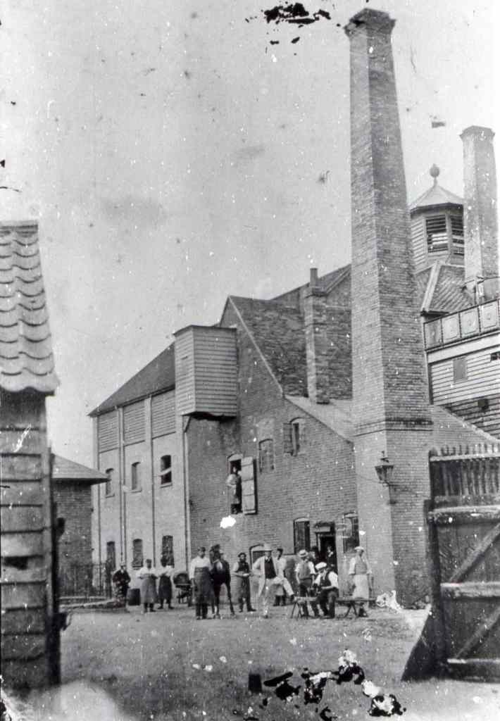 This black and white image shows workers at the brewery in the foreground some seated one holding a horse. all but one wearing aprons. Behind them is a square building with a tall chimney and an octagonal viewing turret behind the chimney. 