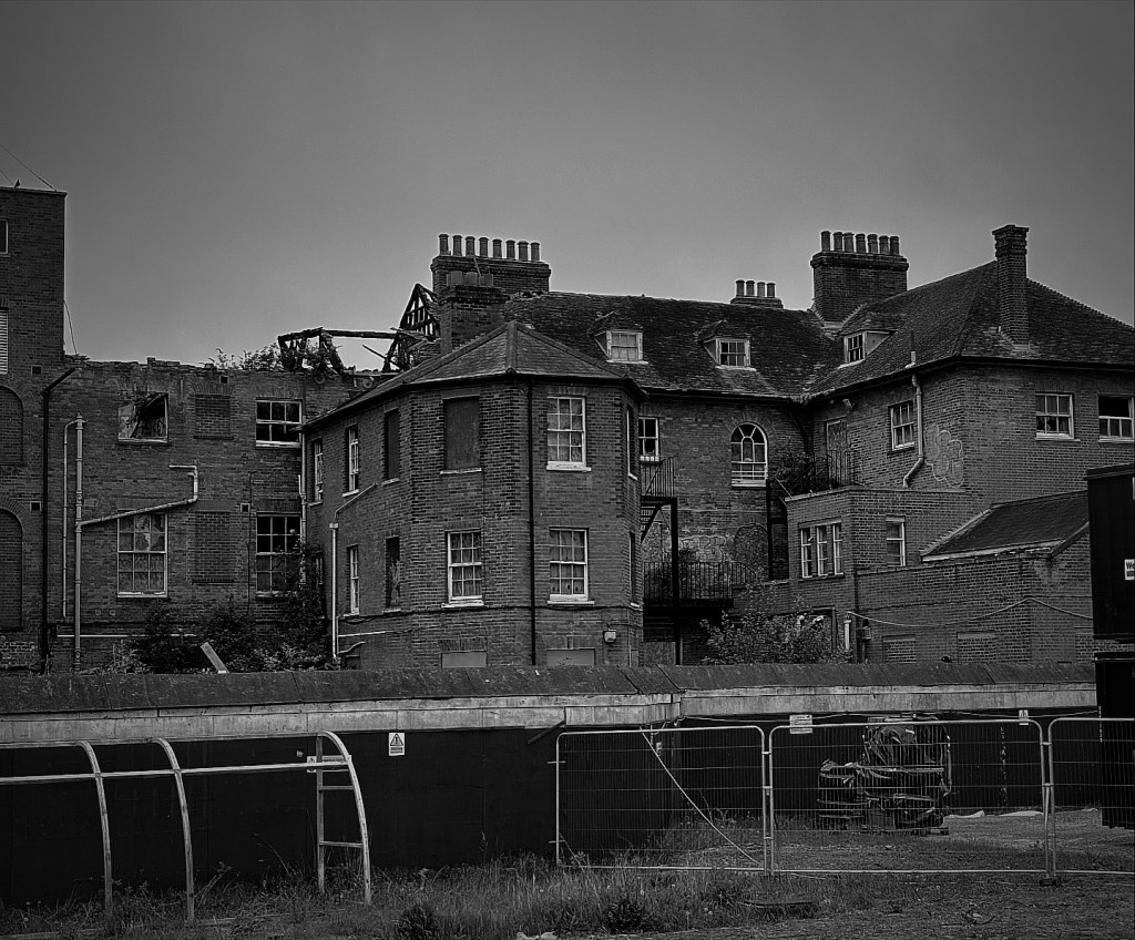 A black and white image of an old Victorian building with an octagonal tower and many chimneys on the left there is no roof and signs of burning at the windows. In front of the building are metal fences and boards to keep the public out. 
