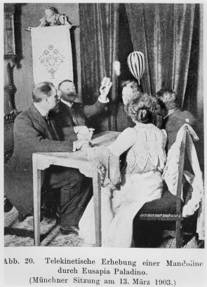 A black and white photo from a newspaper of one woman and four men in late Victorian or Edwardian clothes sitting around a table whilst and object levitates above them. The object is oval with stripes. 