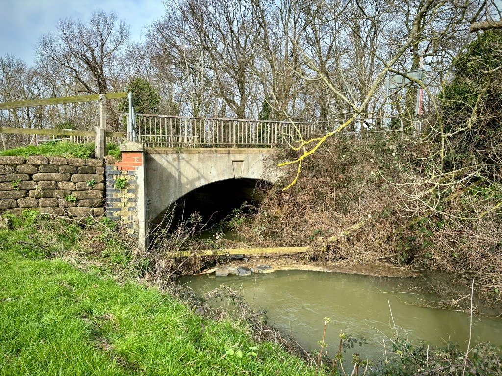 A stone bridge and railings across a wide green but shallow stream. There are winter trees and branches. Above the bridge is a road. To the left the bridge is made of old stones but the main part of the bridge is modern concrete. The sky is blue. 