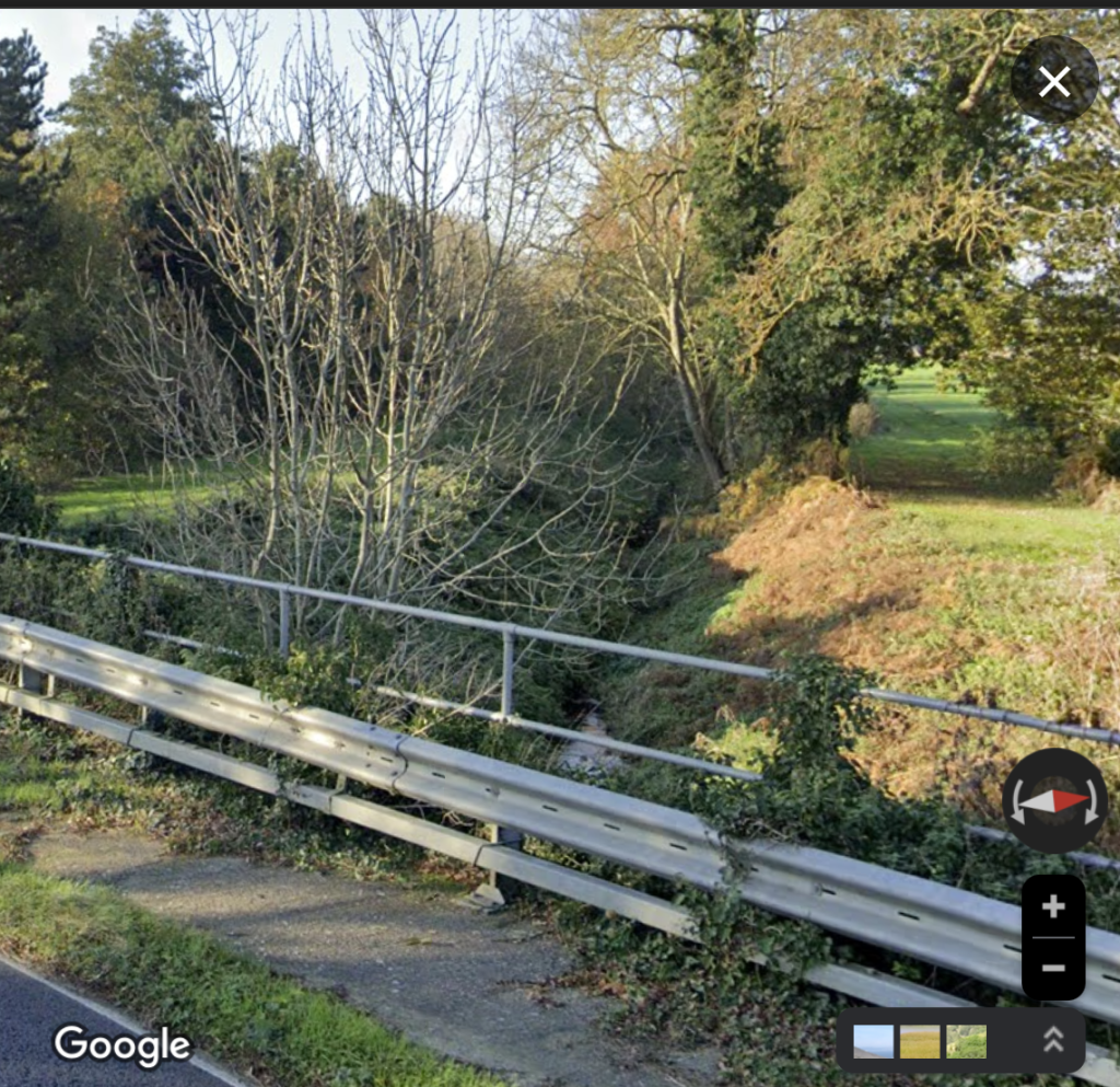 A metal safety railing at the side of a road on a bridge which goes over a narrow ditch of water. A winter tree has fallen to the side of the ditch. The Google logo is on the page.