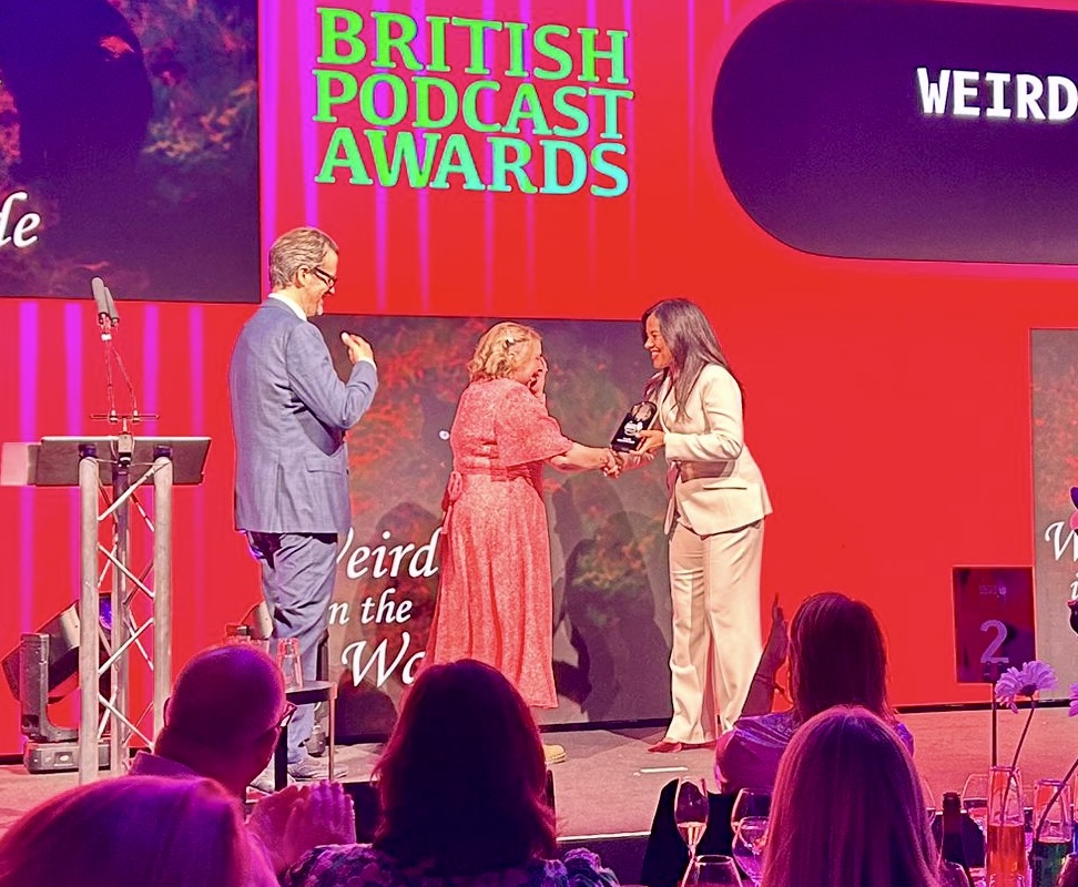 From left to right Marcus Brigstock in a grey suit, me in a pink floaty dress I have blonde wavy hair and I have my hand over my face in shock. Liz Bonnin is wearing a cream suit and is handing me the gold award. The back ground is red with the Weird in the Wade logo on it also.