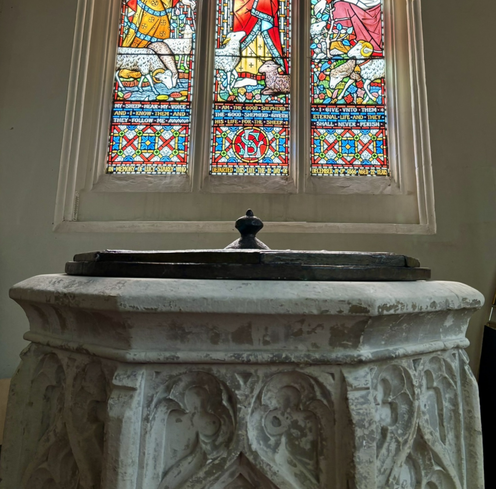 An ornately carved pale stone font with wooden lid. Behind it are bright stained glass windows.