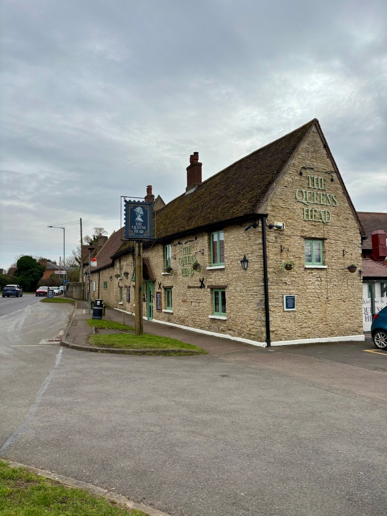 The side and front of a pale beige brick building with mossy roof and large chimneys. The Queens Head is written on the end of the building and the front and the pub sign is penny black stamp. The sky is grey cloud. 