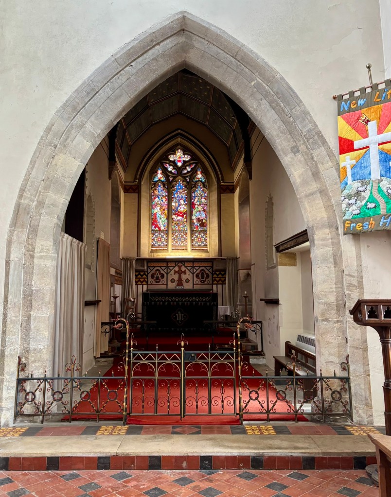 A wider angle of the chancel showing the ornate iron gate at the entrance to the chancel. The archway into the chancel and a banner to the right with white crosses against a sunrise image. The chancel is unusually narrow and dark. 