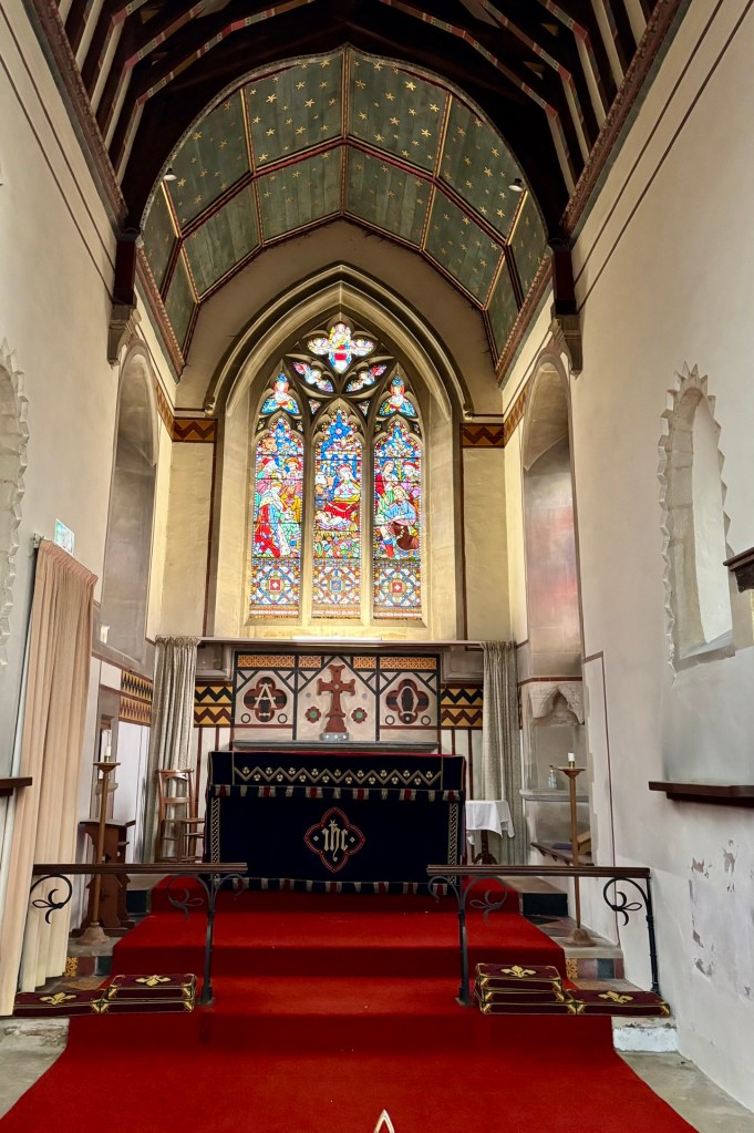 A close up of the chancel with a red carpet and steps leading up to a altar with a black cloth on it which is embroidered in white and red. Behind it are images of the cross and the alpha and omega symbols. There's a stained glass windows and a canopy on the ceiling of gold stars on a blue panels. Then very old wood beams. The walls are white washed and uneven. 