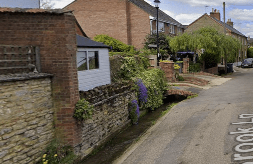 On the left pretty cottages with purple and yellow flowers on their walls in front of the houses is a stream with curved bridges crossing the stream to the houses. Then a road 