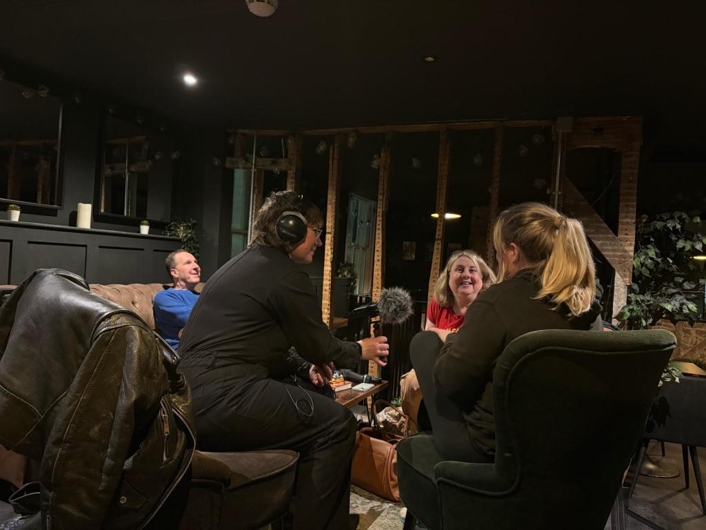 a group of people sitting around a table in the coffeeshop Lucy with headphones and a mic is sitting next to Sarah who has a blonde pony tail and is being interviewed. I am opposite smiling wearing a red t-shirt on the left is Paul wearing a blue top and smiling. The res of the coffee shop is dark. 