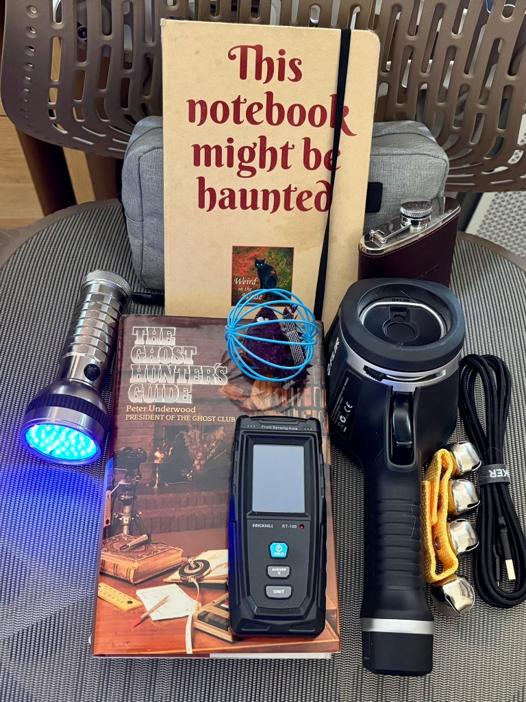 from left to right, a silver torch with a purple light, a book called The Ghost Hunters Guide by Peter Underwood, a cat ball toy, a black hand held EMF reader, a large thermal imaging camera, a set of sleigh bells, a hip flask and a This Notebook Might Be Haunted weird in the wade note book. 
