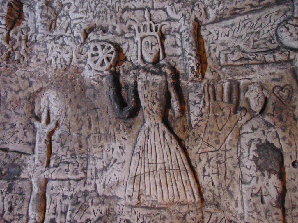 A carving of a woman in a long striped skirt with a cross on it holding a wheel. She has a large crown on her head. 