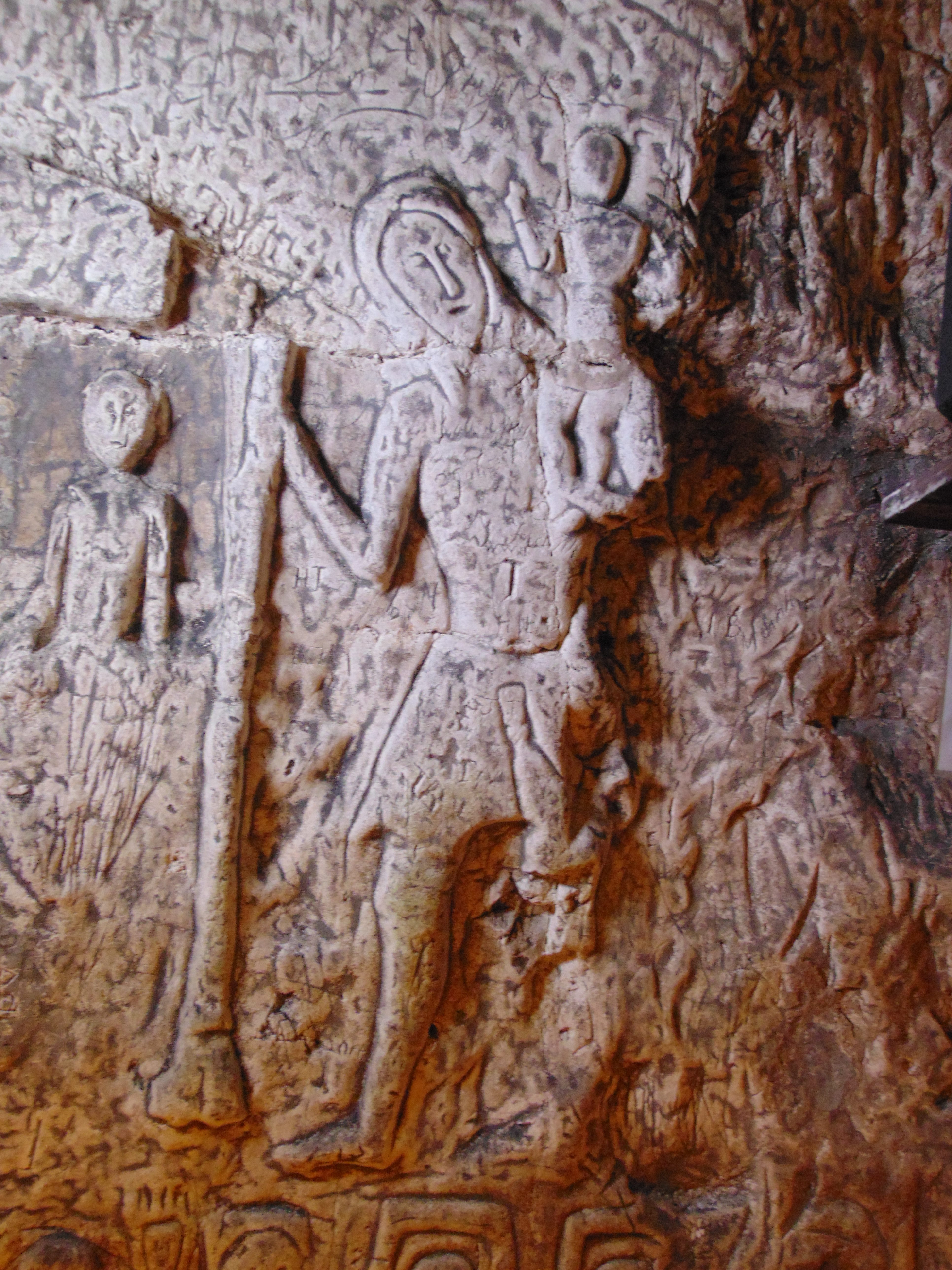 A tall deeply carved figure holding a staff in their left hand and they are carrying a child on their right shoulder. 