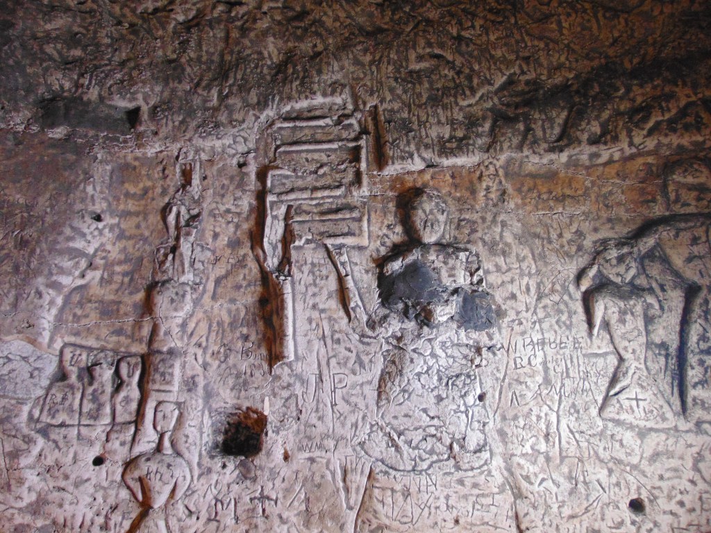 A carving of a central figure with a deeply carved head but little in the way of features. He holds a griddle or grill in his hand.