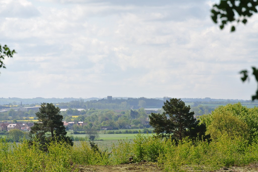 A view across a large flat valley to higher ground in the distance. There are trees and yellow fields and a few houses. On the higher ground in the distance there is a water tower. 
