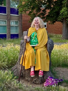 A woman wearing a green dress and yellow cloak sitting on a tree stump and laughing