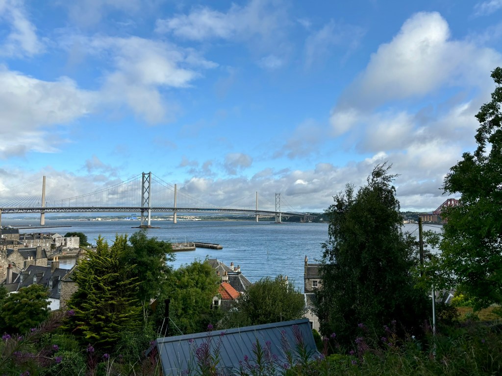 In the foreground there are rooves and small grey stone cottages amongst trees. In the middle distance is a silver bridge across the firth of forth. On the extreme right peaking though the trees is the forth rail bridge. In the distance are the hills of fife. 