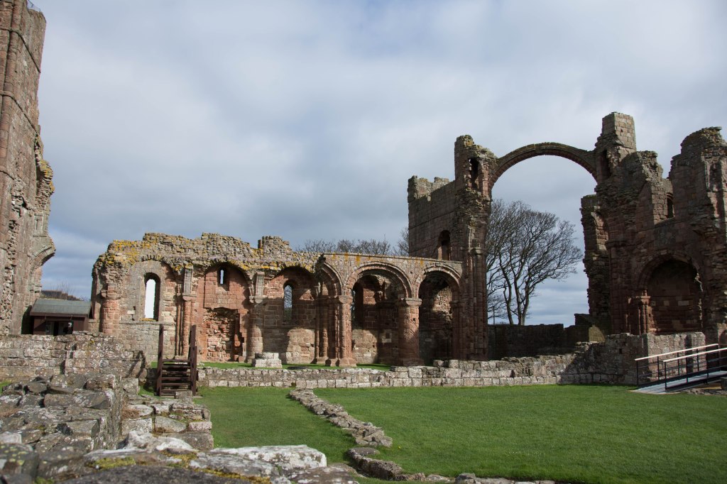 Ruins of Lindisfarne abbey including a greay arch with a winter tree behind it and a collinade of pillars in a warm pink stone. 