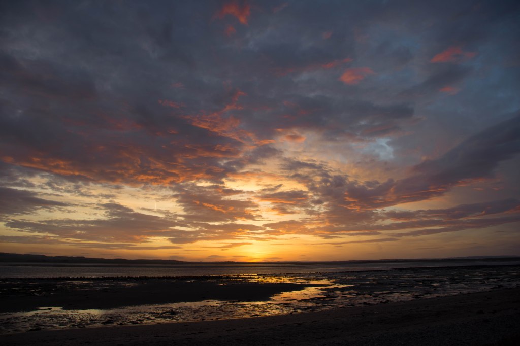 A snake of sea across the sand and rocks turned gold by the rising sun which is catching the bottom of clouds turning them from grey to deep gold and red.